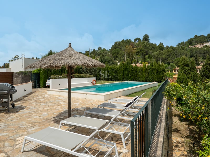 Piscine rectangulaire entourée d'une terrasse en pierre, chaises longues blanches et parasol en paille.