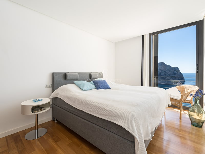 Contemporary bedroom with a gray upholstered bed, wooden floors, and a floor-to-ceiling window overlooking the sea.