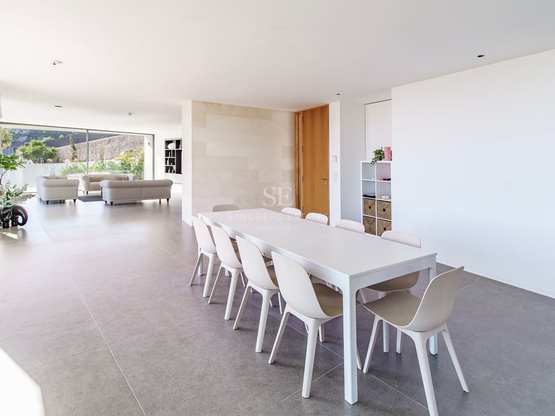 Salle à manger minimaliste et lumineuse avec table et chaises blanches, s'ouvrant sur un grand salon vitré.