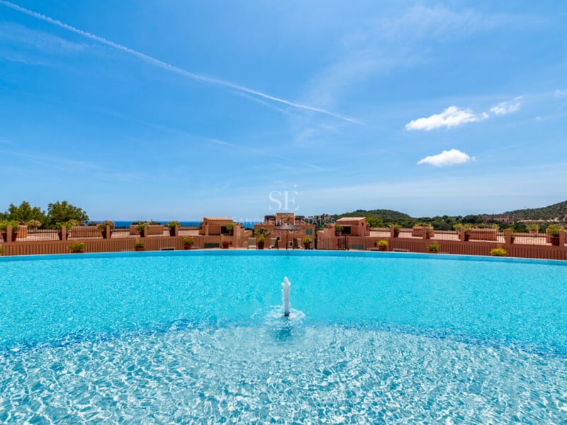 Grande piscine incurvée avec fontaine centrale et vue panoramique sur les bâtiments méditerranéens et la mer.