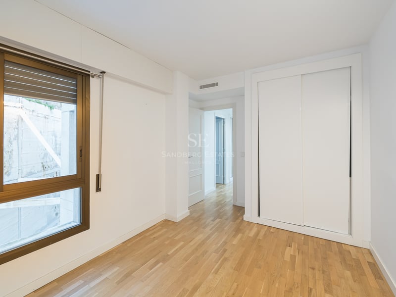 Tomt rum med ljust trägolv, en vit inbyggd garderob och ett stort fönster med bronsfärgad ram.