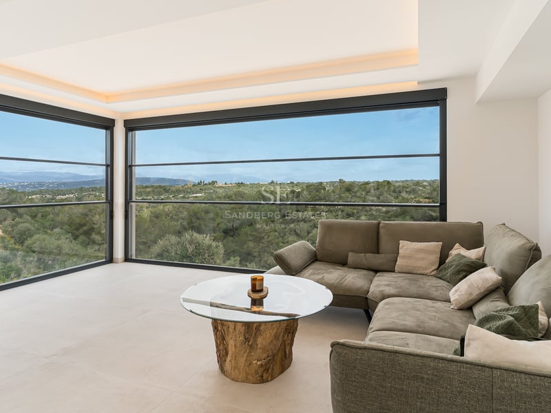Zeitgenössisches Wohnzimmer mit grauem Sofa, Holz-Couchtisch und großen Fenstern mit Blick auf grüne Landschaft.