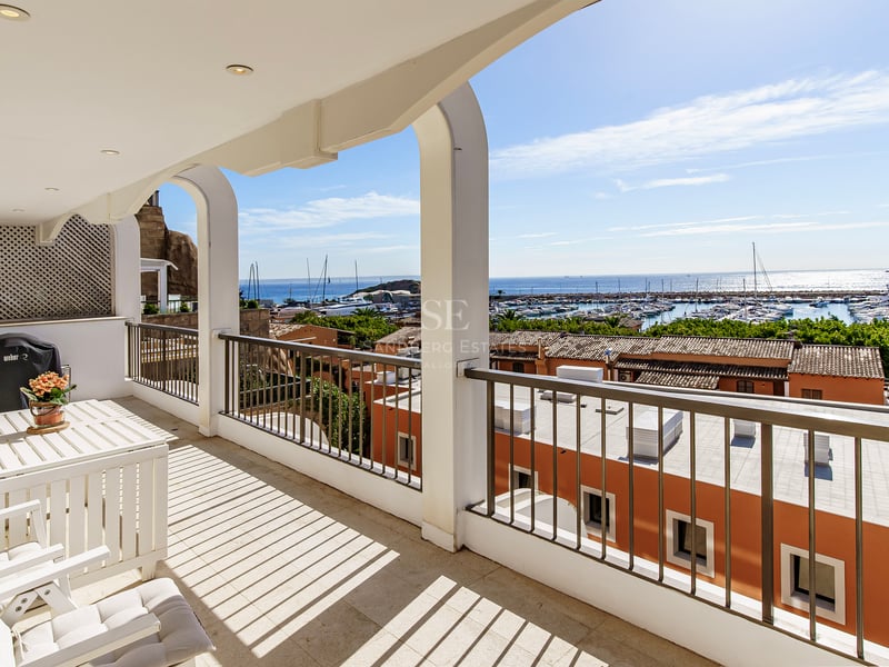 White arched balcony with outdoor furniture overlooking a harbor with boats and blue sea under a clear sky.