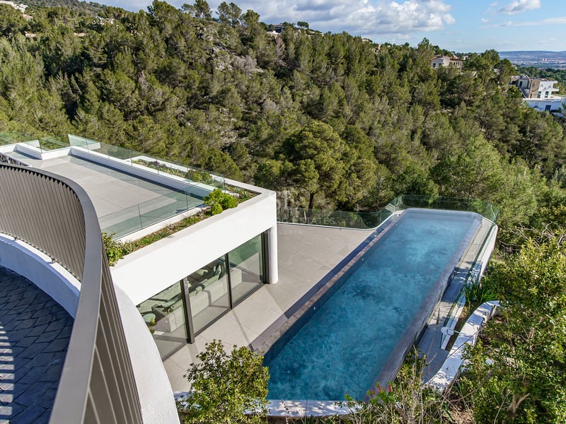Vue en plongée d'une piscine à débordement d'une villa moderne surplombant une forêt verdoyante sous un ciel dégagé.