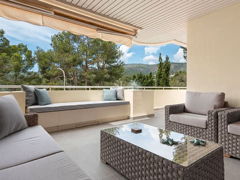 Covered terrace with grey wicker lounge furniture, a built-in bench, and views of pine trees and mountains.