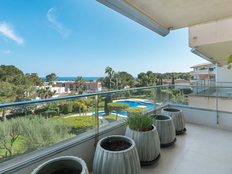 Modern balcony with glass railings overlooking lush green gardens and the blue sea under a clear sky.