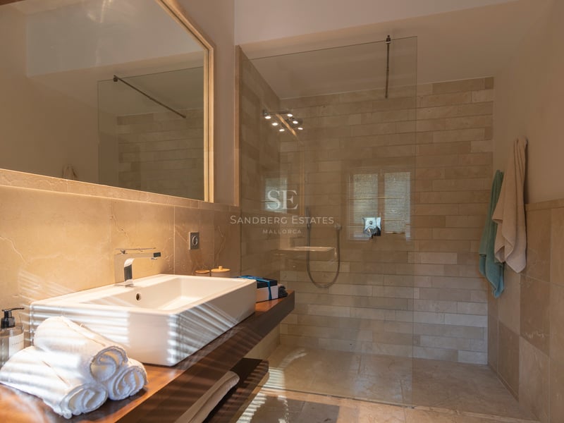 Modernt badrum med duschvägg i glas, kakel i natursten och kommod i mörkt trä med ovanpåliggande tvättställ.