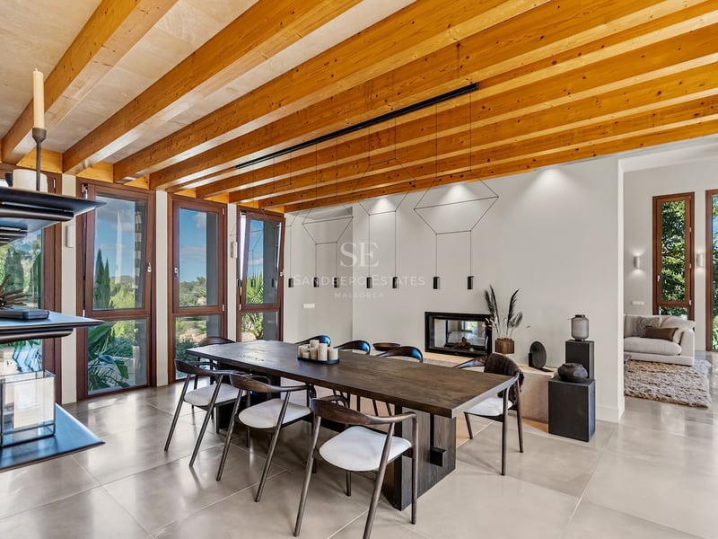 Grande table à manger en bois sous un plafond à poutres avec des baies vitrées donnant sur la nature.