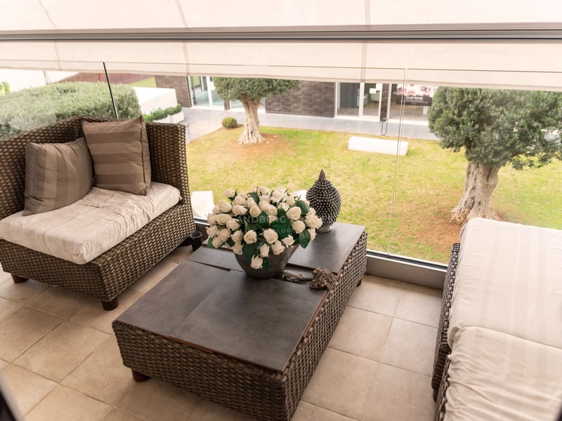 Überdachte Terrasse mit Rattan-Loungemöbeln, Glasgeländer und Blick auf einen Garten mit Olivenbäumen.