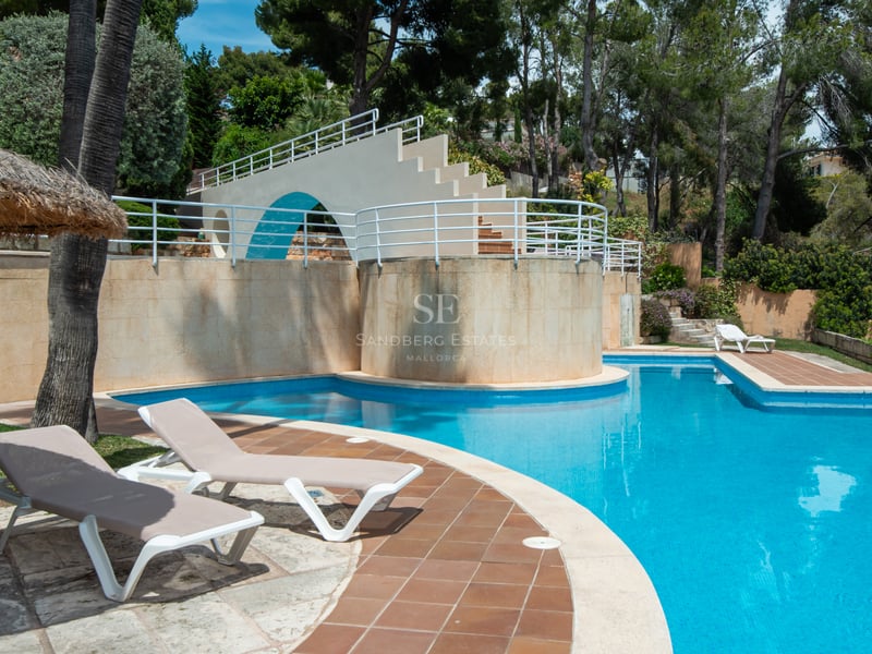 Large blue tiled swimming pool with sun loungers, a straw umbrella, and a decorative white bridge structure in a garden.