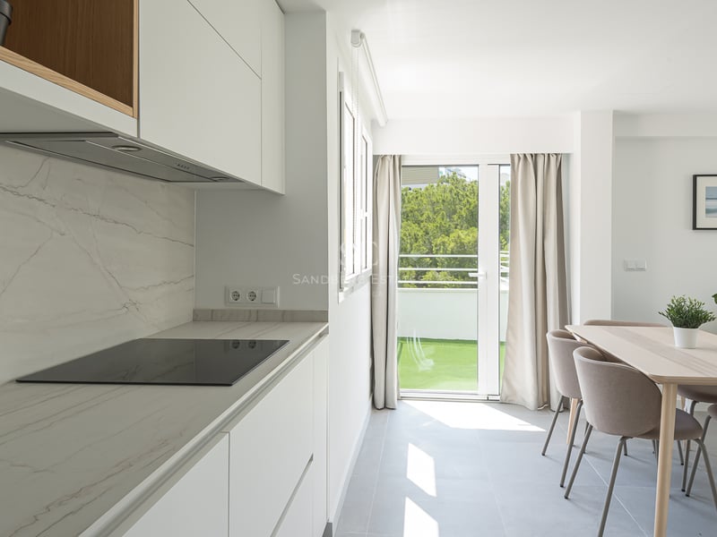 Cuisine blanche moderne avec plan de travail en marbre, salle à manger en bois et porte de balcon.