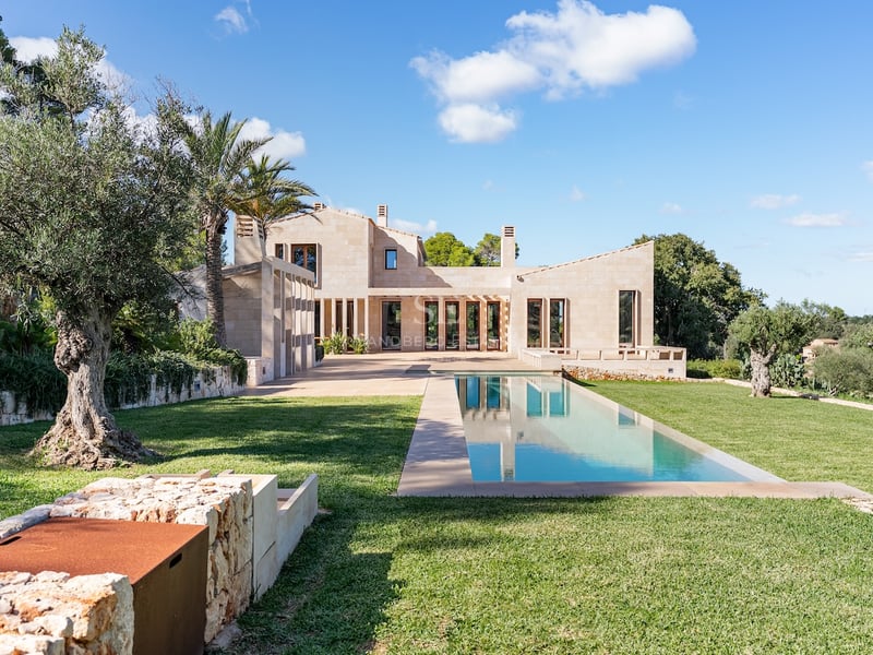 Villa moderne en pierre avec une longue piscine rectangulaire, pelouse verte et oliviers centenaires sous un ciel bleu.