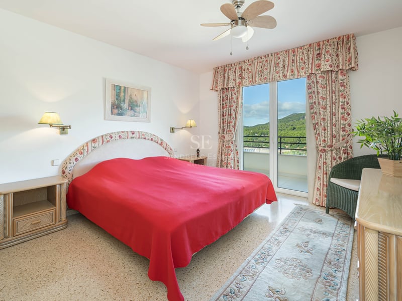Bright bedroom featuring a red bedspread, floral curtains, and glass doors opening to a balcony with green hill views.