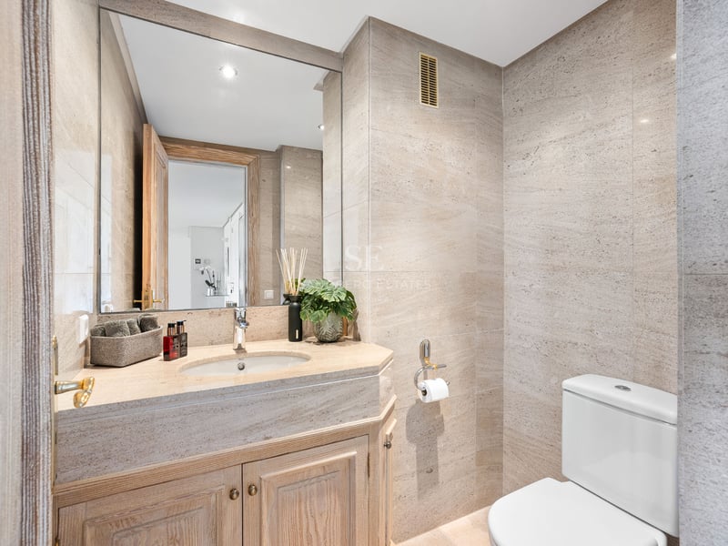 Salle de bains moderne avec carrelage en pierre beige, grand miroir et meuble sous vasque en bois.