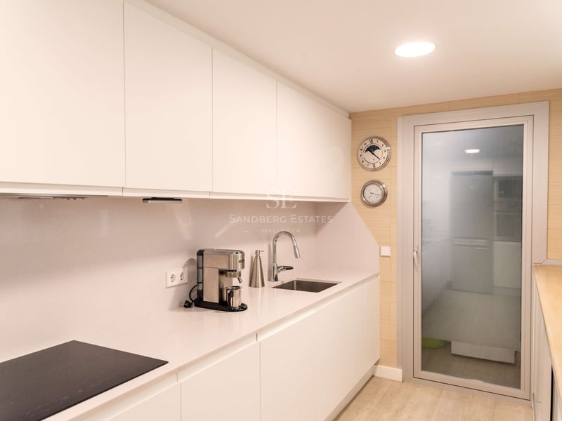 Moderne weiße Küche mit grifflosen Schränken, Quarz-Arbeitsplatte und Glastür zum Außenbereich.