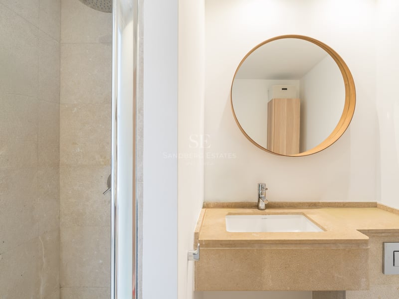 Modernt badrum med tvättställ i beige sten, rund träspegel och duschkabin i glas.