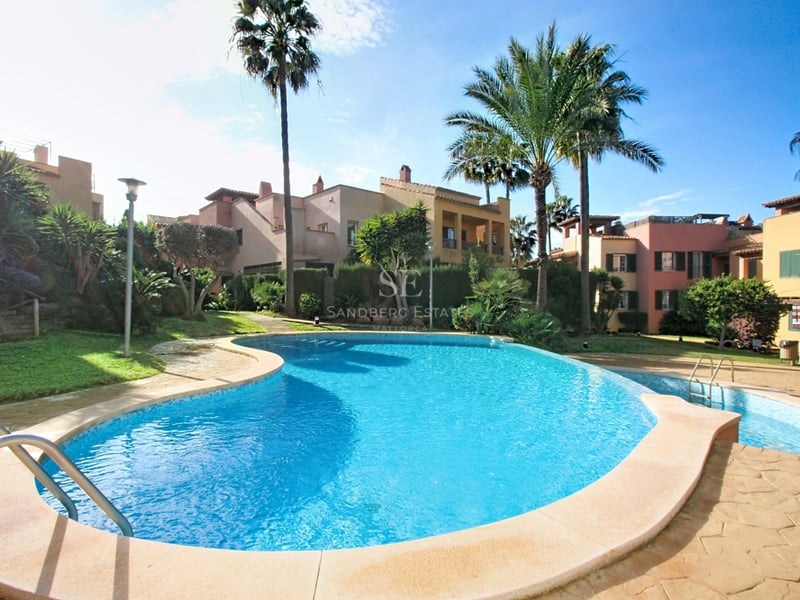 Oregelbundet formad pool omgiven av tropiska trädgårdar och byggnader i medelhavsstil i dagsljus.