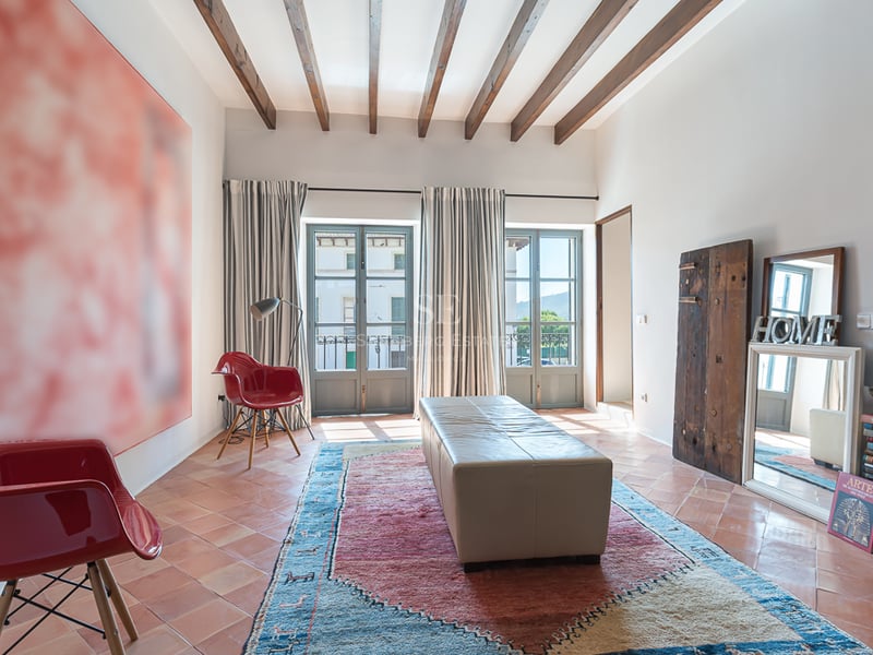 Espace de vie avec sols en terre cuite, poutres en bois sombre, chaises rouges et deux balcons français lumineux.