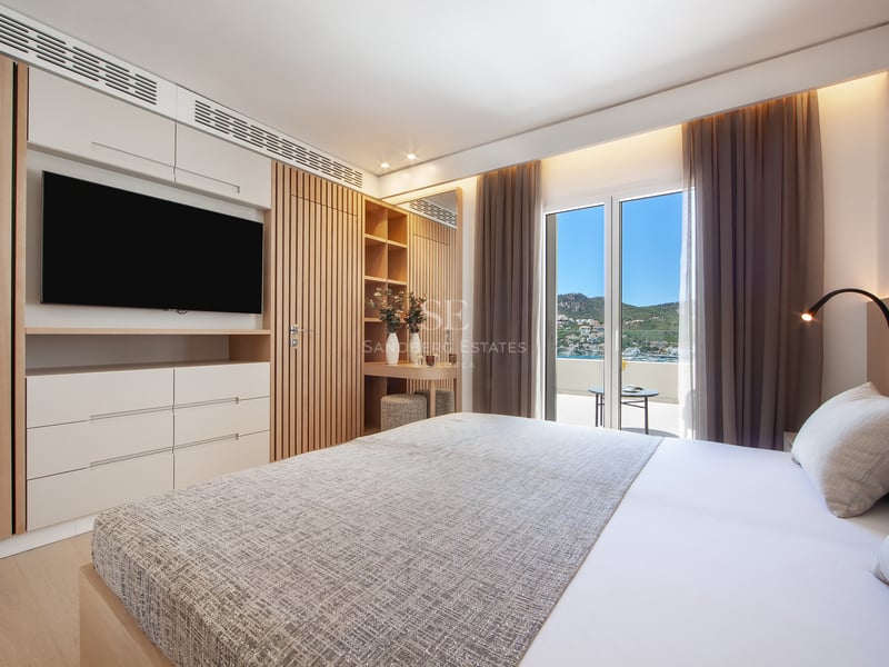 Chambre lumineuse avec grand lit, boiseries, TV et balcon offrant une vue sur la côte et les montagnes.