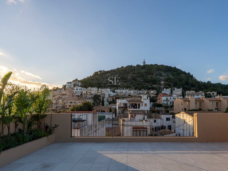 Grande terrasse carrelée avec garde-corps métallique offrant une vue sur un village méditerranéen et une colline au coucher du soleil.