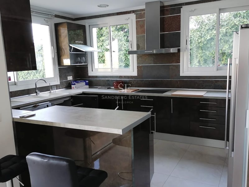 Cuisine moderne en L avec placards sombres brillants, plan de travail gris et fenêtres donnant sur le jardin.