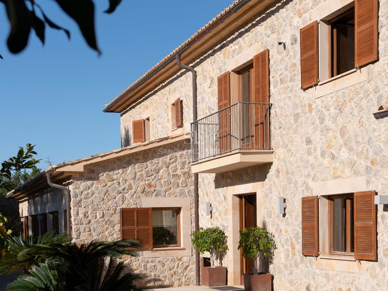 Exteriör av en tvåvånings stenvilla med fönsterluckor i trä och en balkong under en klarblå himmel.