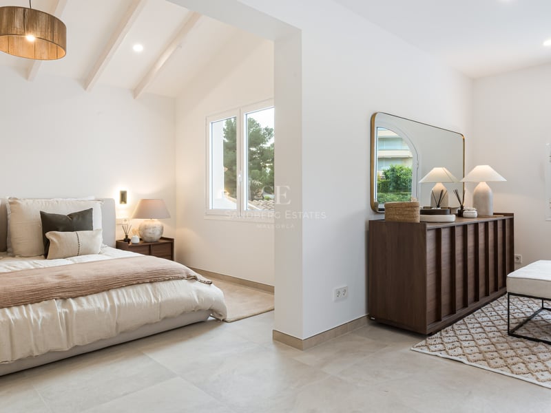 Chambre parentale moderne avec plafonds à poutres blanches, lit king-size, commode en bois sombre et lumière naturelle.