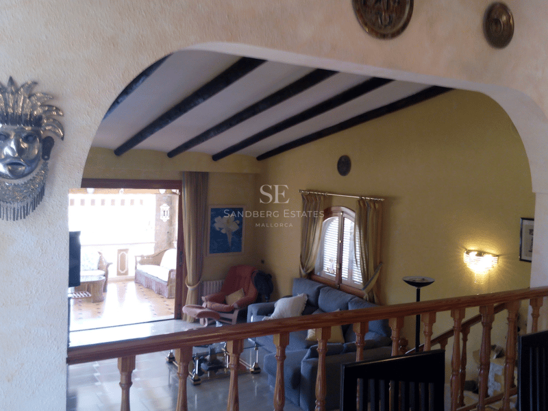 Salón acogedor visto a través de un arco con vigas de madera oscura y acceso a terraza sombreada.