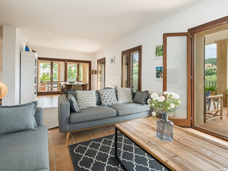 Canapé gris moderne et table basse en bois dans un salon lumineux avec sol en terre cuite et grandes baies vitrées.