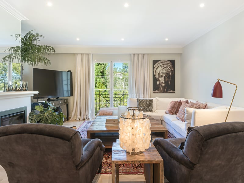 Bright living room with white L-shaped sofa, dark armchairs, wooden coffee table, and balcony overlooking greenery.