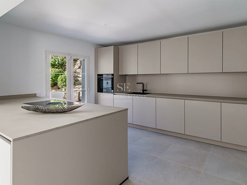 Samtida kök med sandfärgade handtagslösa skåp, central köksö och utgång till trädgård.