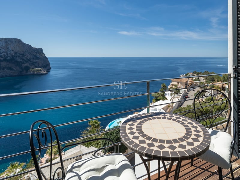 A small terrace with a mosaic table and iron chairs overlooking a deep blue Mediterranean bay and rugged cliffs.