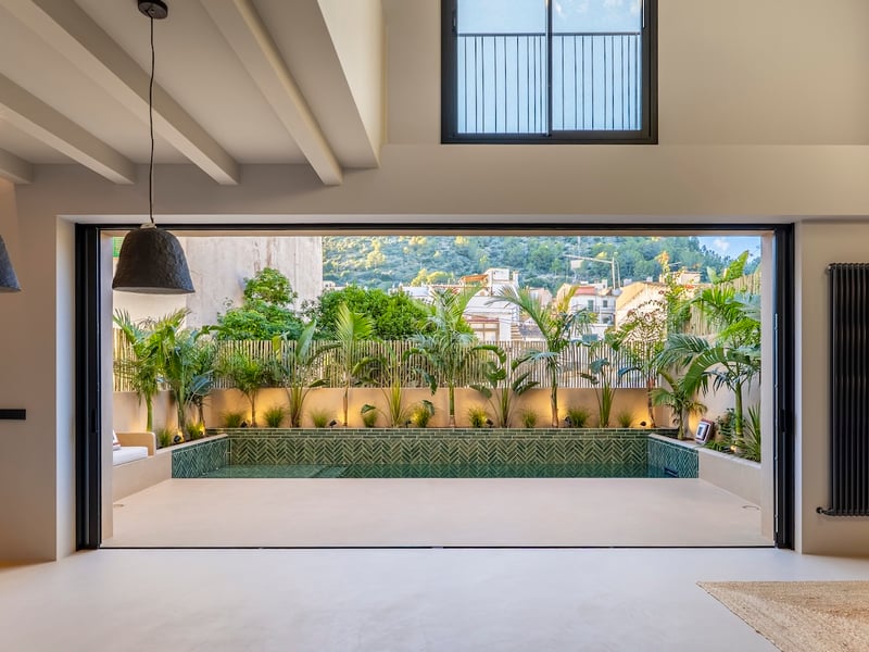 Vue intérieure par une grande baie vitrée vers une piscine privée entourée de plantes tropicales et carrelage vert.
