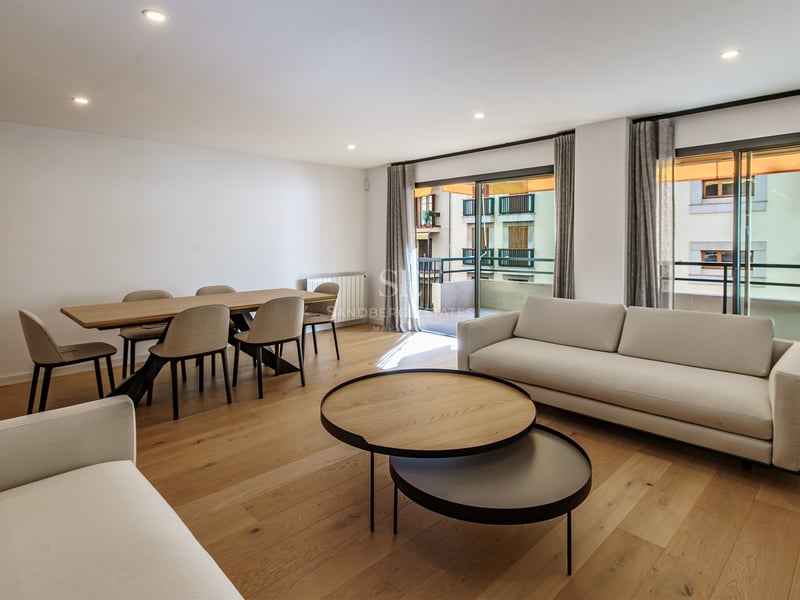 Salon moderne avec canapés blancs, tables basses en bois, coin repas et grandes baies vitrées ouvrant sur un balcon.