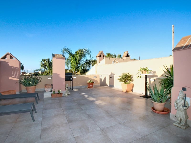 Stor takterrass med beige klinker, rosa putsade väggar, solstolar, krukväxter och klarblå himmel.