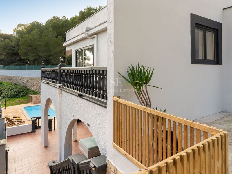 Villa blanche à plusieurs niveaux avec balustrades noires et en bois, surplombant une piscine turquoise et la forêt.