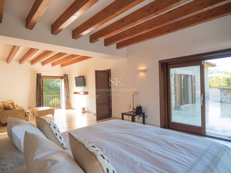 Rymligt sovrum med högt i tak, takbjälkar i trä, stengolv och skjutdörrar i glas mot terrassen.