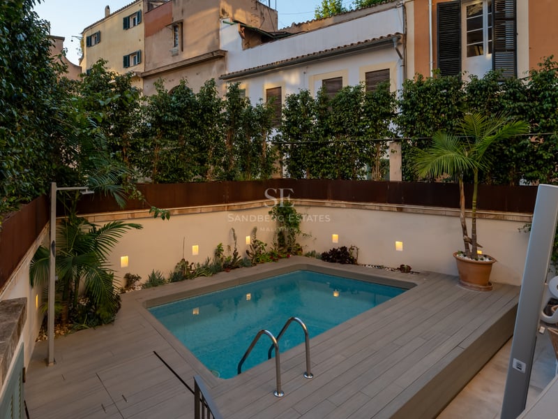 Petite piscine rectangulaire dans une cour avec un deck en bois, entourée de hautes haies vertes et d'appliques murales.