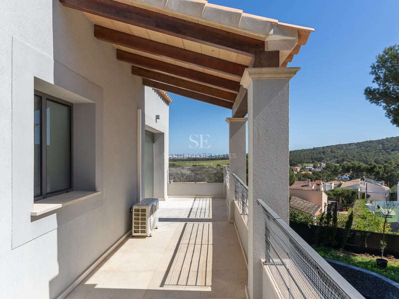 Lång övertäckt balkong med träbjälkar, vita väggar och metallräcke med utsikt över en grönskande dal.