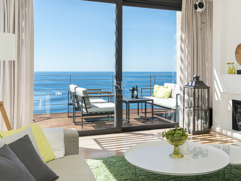 Contemporary living room with white decor, fireplace, and floor-to-ceiling glass doors opening to a sea-facing terrace.