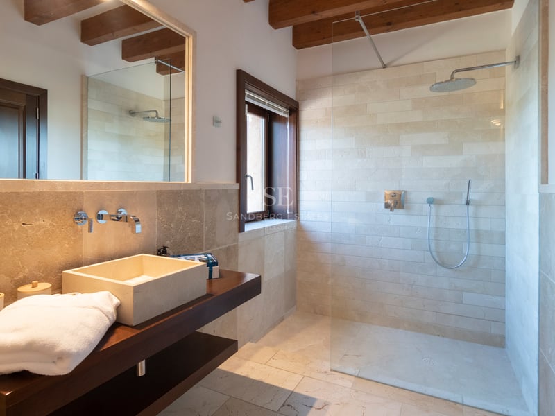 Modernt badrum med naturstensväggar, walk-in takdusch och synliga takbjälkar i trä.