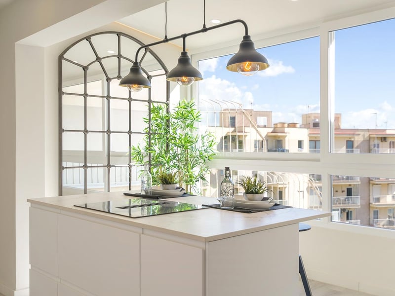 Weiße Kücheninsel mit Induktionskochfeld, Industrielampen und großen Fenstern mit Blick auf Stadtgebäude.