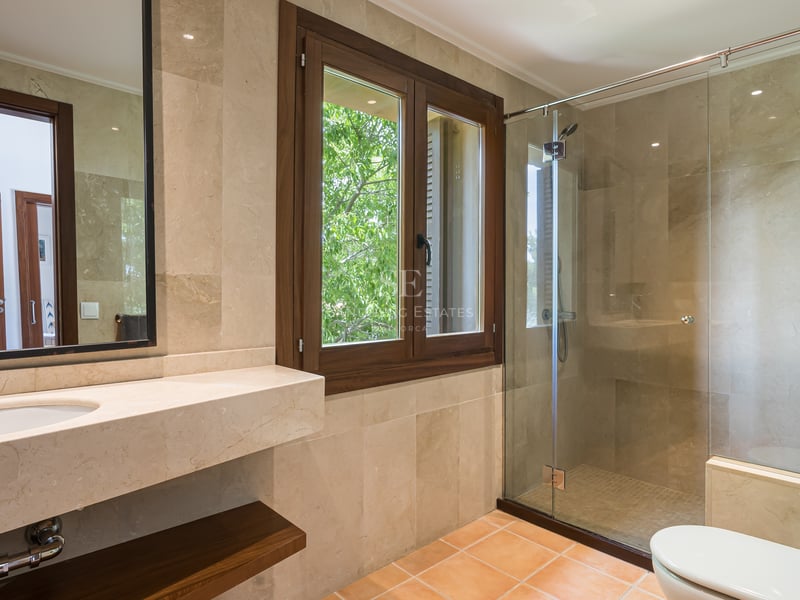 Salle de bain avec douche à l'italienne vitrée, carrelage en pierre beige et fenêtre en bois sur le jardin.