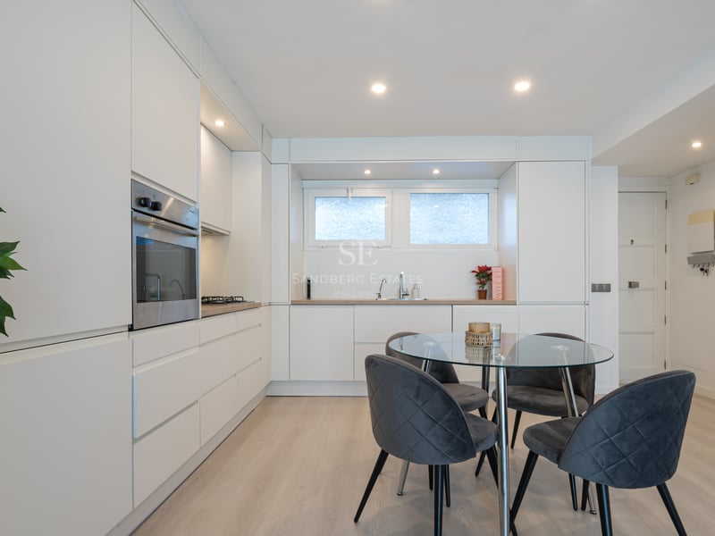 Clean white minimalist kitchen featuring handleless cabinetry, glass dining table, and grey velvet chairs.