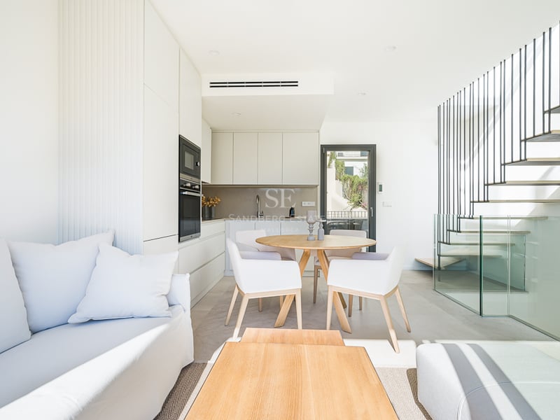 Bright open-plan room with white sofa, wooden dining set, integrated kitchen, and a floating staircase with glass railing.
