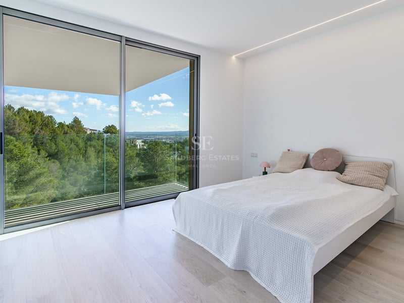 Chambre minimaliste lumineuse avec de grandes baies vitrées ouvrant sur un balcon avec vue sur les collines verdoyantes.