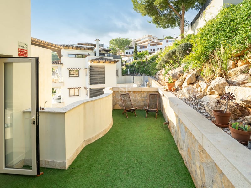 Solig terrass med grönt konstgräs, två loungestolar i trä och en naturstensvägg.