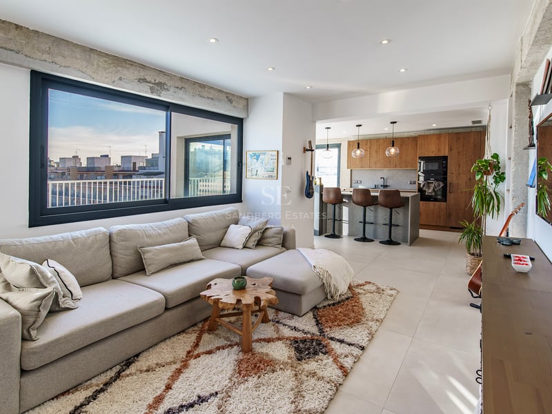 Salon moderne et lumineux avec un grand canapé d'angle gris, un tapis texturé et une cuisine ouverte avec vue sur la ville.