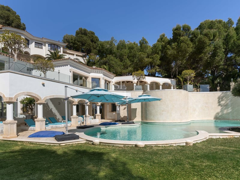 Geschwungener Pool mit türkisfarbenen Schirmen neben einer weißen Mittelmeervilla, umgeben von Pinien.
