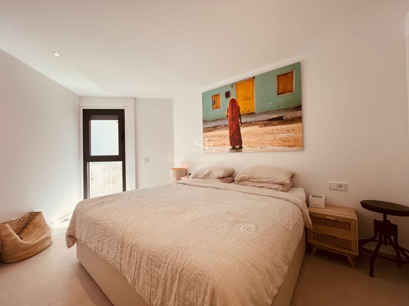 Chambre minimaliste avec grand lit, parure de lit beige, table de chevet en bois et grand tableau coloré.